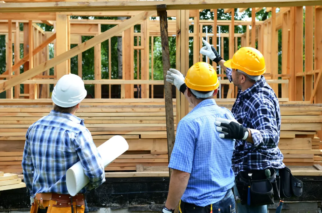 Three construction workers in hard hats discussing a wooden frame structure, with one holding blueprints.
