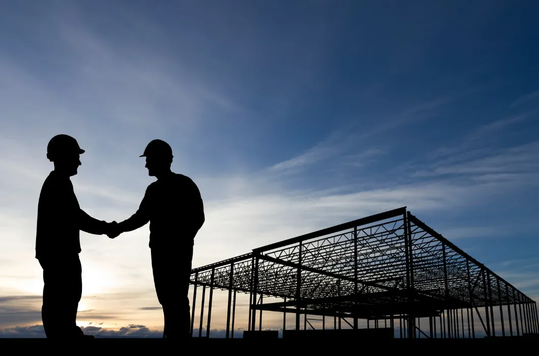 Two construction workers in silhouette shaking hands at a building site against a sunset sky.