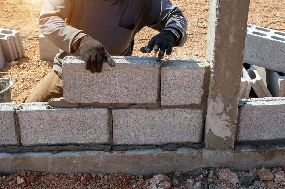 Worker laying concrete blocks with gloves, using a yellow string for alignment at a construction site.