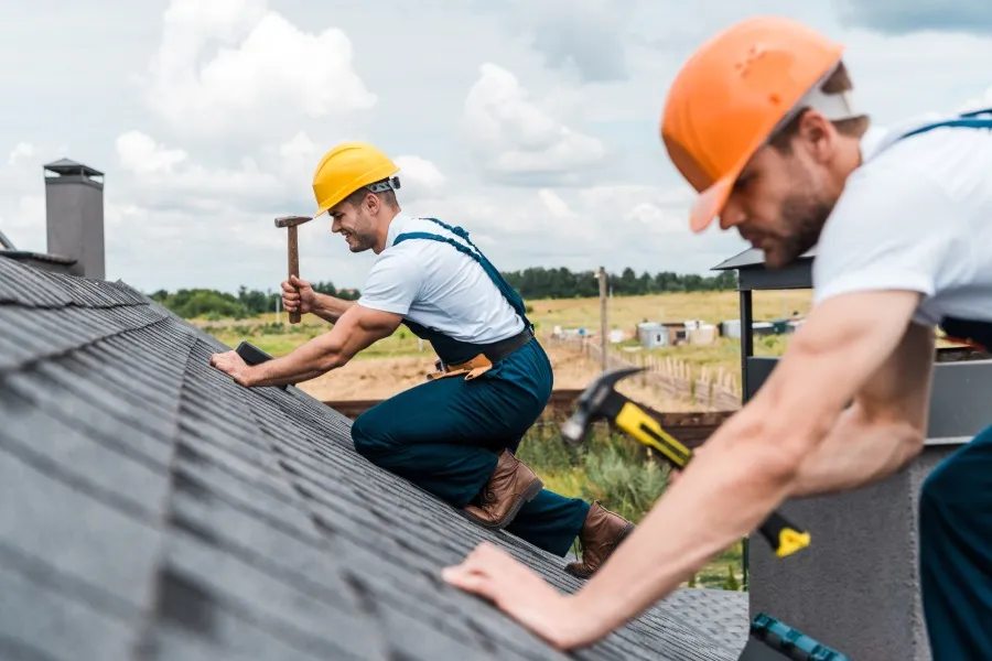 Two workers in hard hats and overalls repairing a roof, with one using a hammer, under a cloudy sky.