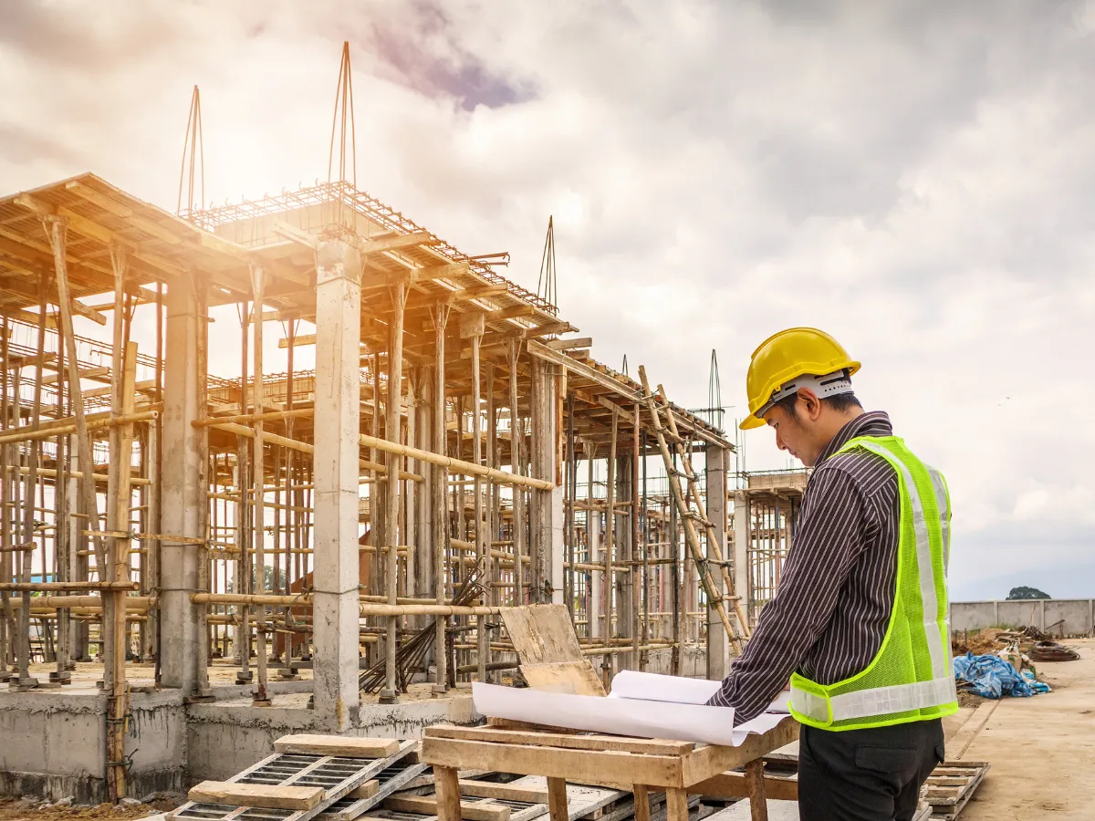 Construction worker inspects blueprints on site with a partially built structure and scaffolding in the background.