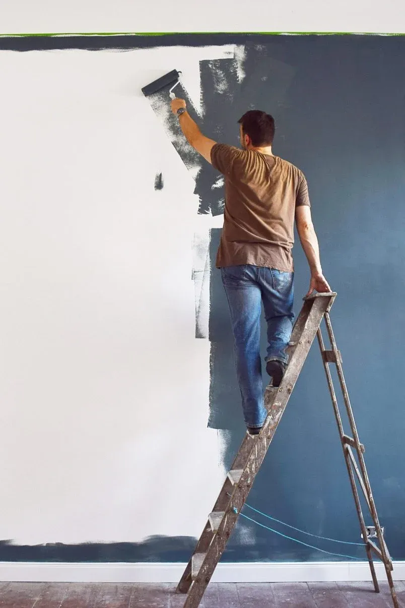 Person standing on a ladder painting a wall dark blue with a roller.