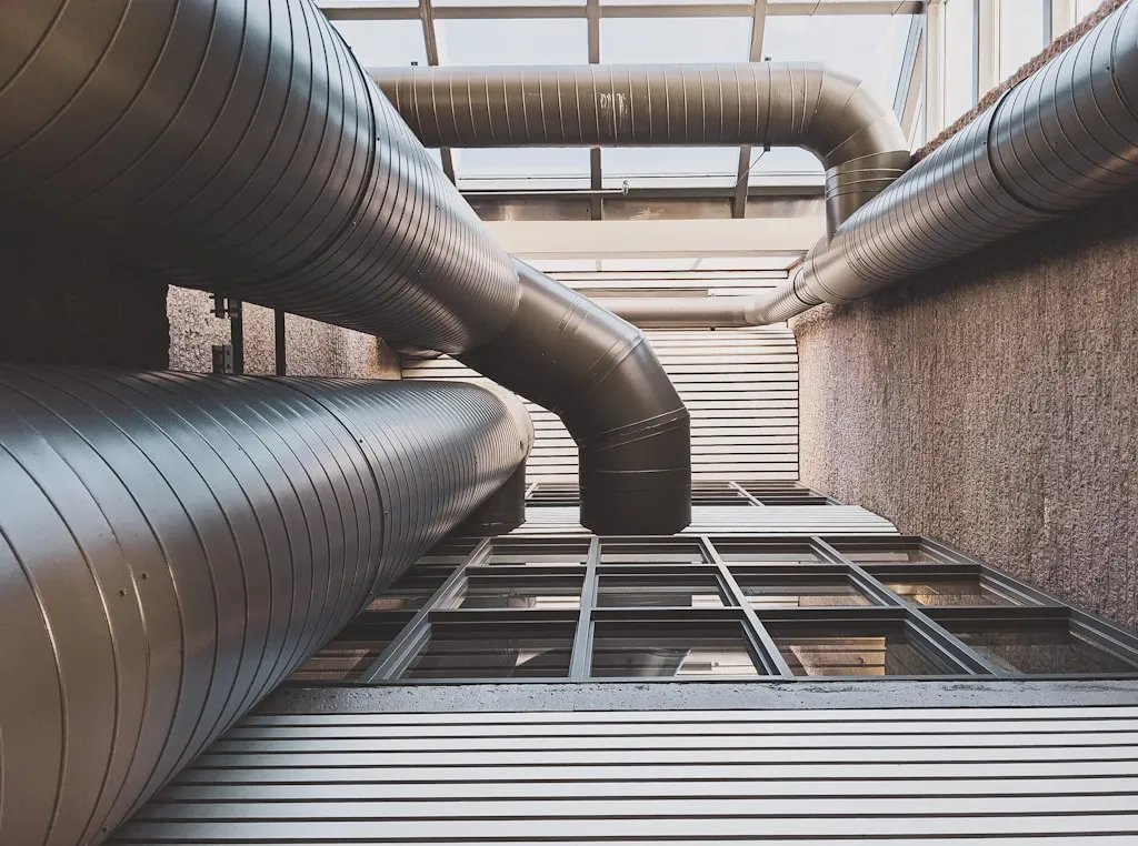 View from below of industrial vents and pipes against a modern building facade with a windowed roof.