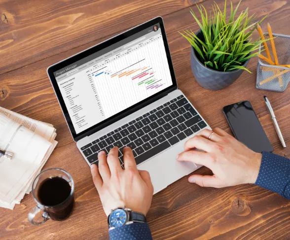 Hands typing on a laptop displaying a Gantt chart, next to a phone, coffee cup, plant, and notebook on a wooden table.
